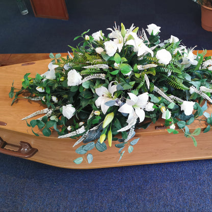 Artificial floral arrangement of white lilies and green foliage displayed on a light wood casket inside a room with blue carpeting.
