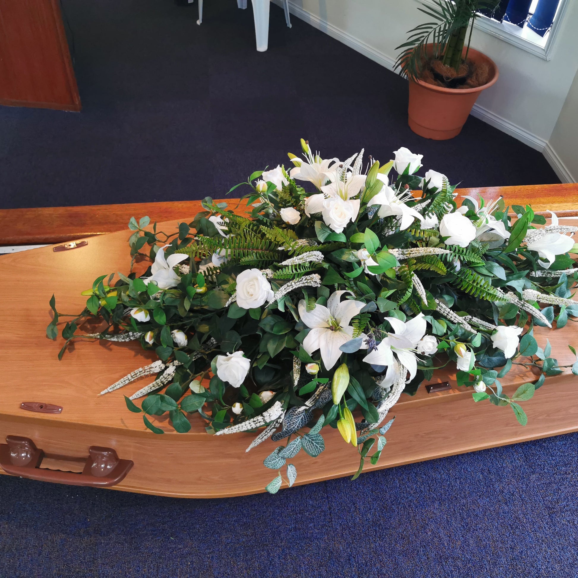 A wooden casket with a large artificial  white lily and green foliage floral arrangement placed on top, photographed indoors on blue carpet.