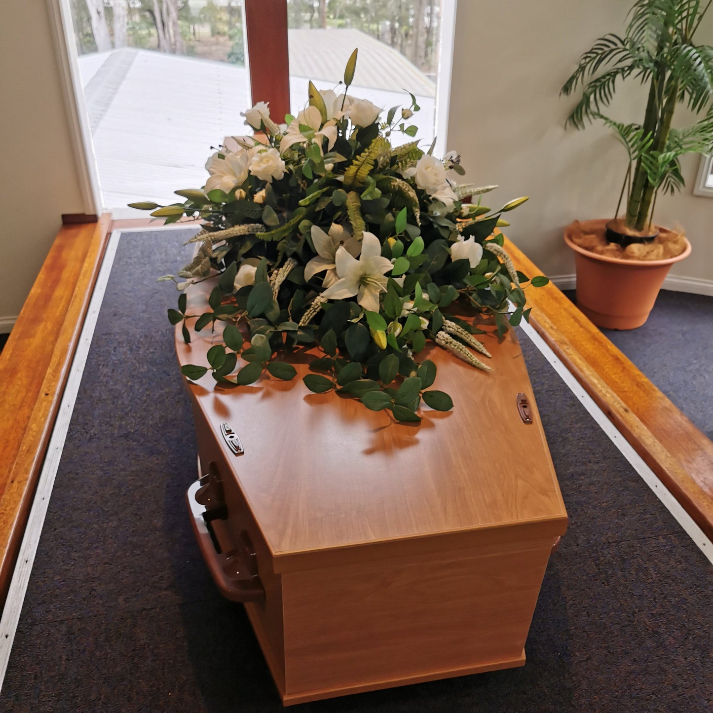 A wooden casket with a floral arrangement of white lilies and green foliage, displayed on a dark carpeted aisle in an indoor memorial or funeral setting.