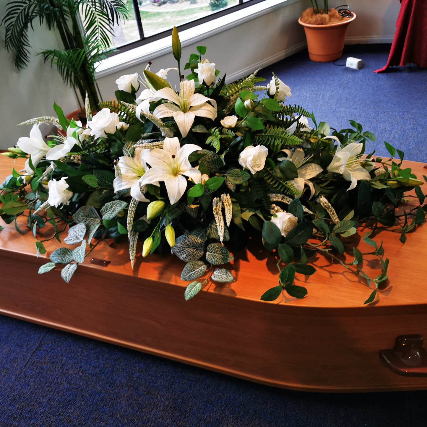 A large artificial floral arrangement with white lilies and green foliage draped across a polished wooden casket on a blue carpeted floor in an indoor venue.