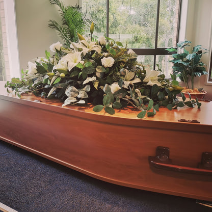A polished reddish-brown wooden casket with metal handles, displayed indoors on a blue carpet in front of large windows. A large floral arrangement with artificial white flowers and green foliage rests across the top of the casket.