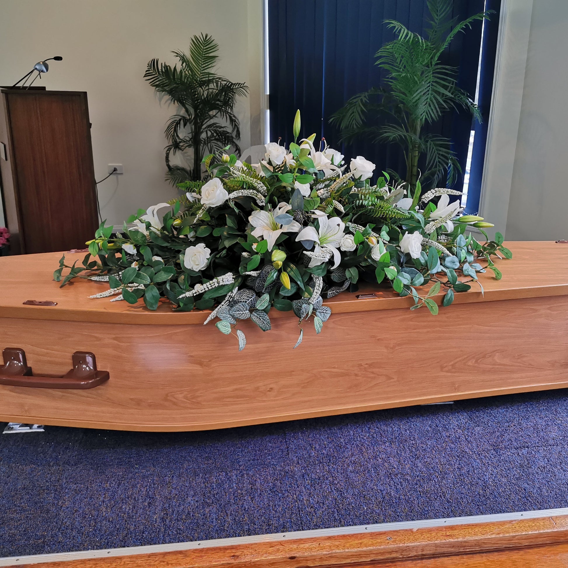 A polished light brown wooden casket with dark brown metal handles, decorated with Everlasting Funeral Flowers, a large white and green floral arrangement, set on a blue carpeted stage.