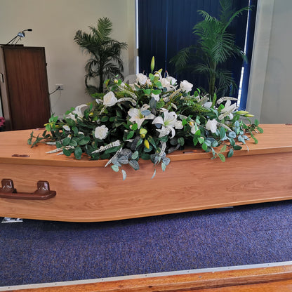 A polished light brown wooden casket with dark brown metal handles, decorated with Everlasting Funeral Flowers, a large white and green floral arrangement, set on a blue carpeted stage.
