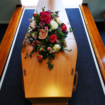 A polished light brown wooden casket with a artificial floral arrangement of fuchsia, pink, and white roses on top, positioned on a dark blue carpeted aisle between wooden pews.