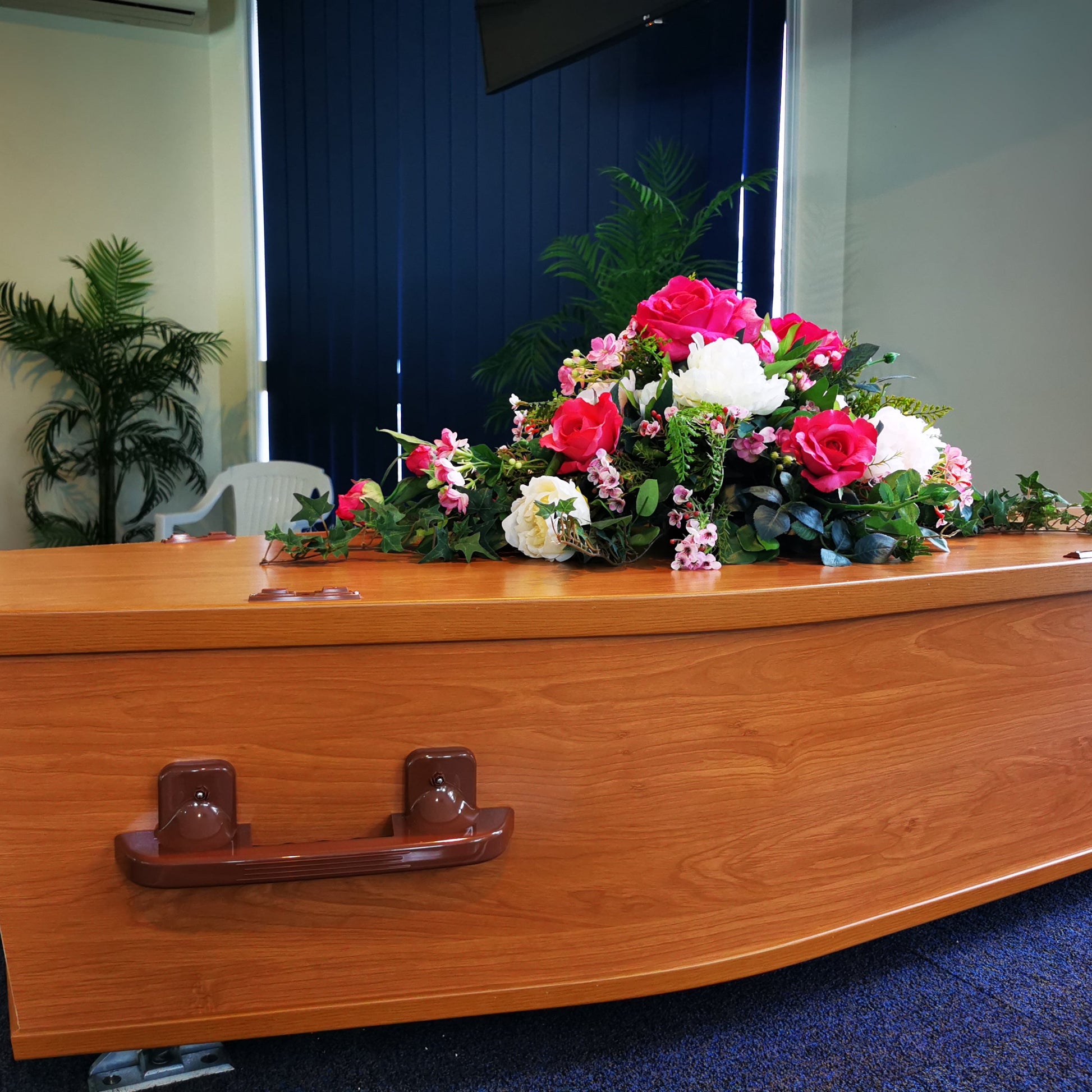 A polished wooden casket with a artificial floral arrangement of pink, fuchsia, and white roses on top, set in an indoor room with blue carpet and a dark blue door in the background.