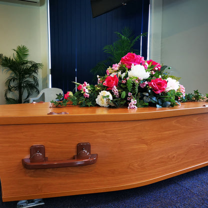 A polished wooden casket with a artificial floral arrangement of pink, fuchsia, and white roses on top, set in an indoor room with blue carpet and a dark blue door in the background.