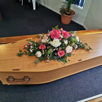 A light natural wood casket with metal handles, decorated with a pink, white, and green artificial floral arrangement, resting on a blue carpeted floor in an indoor setting.