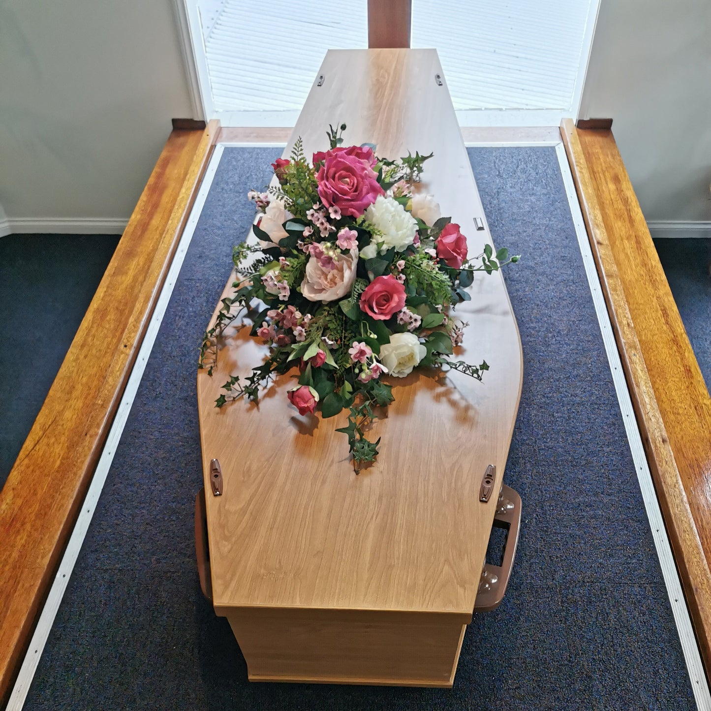 A light natural wood coffin-shaped box or casket with a artificial floral arrangement of pink, red, and white roses on top, photographed indoors on a dark blue carpeted surface.
