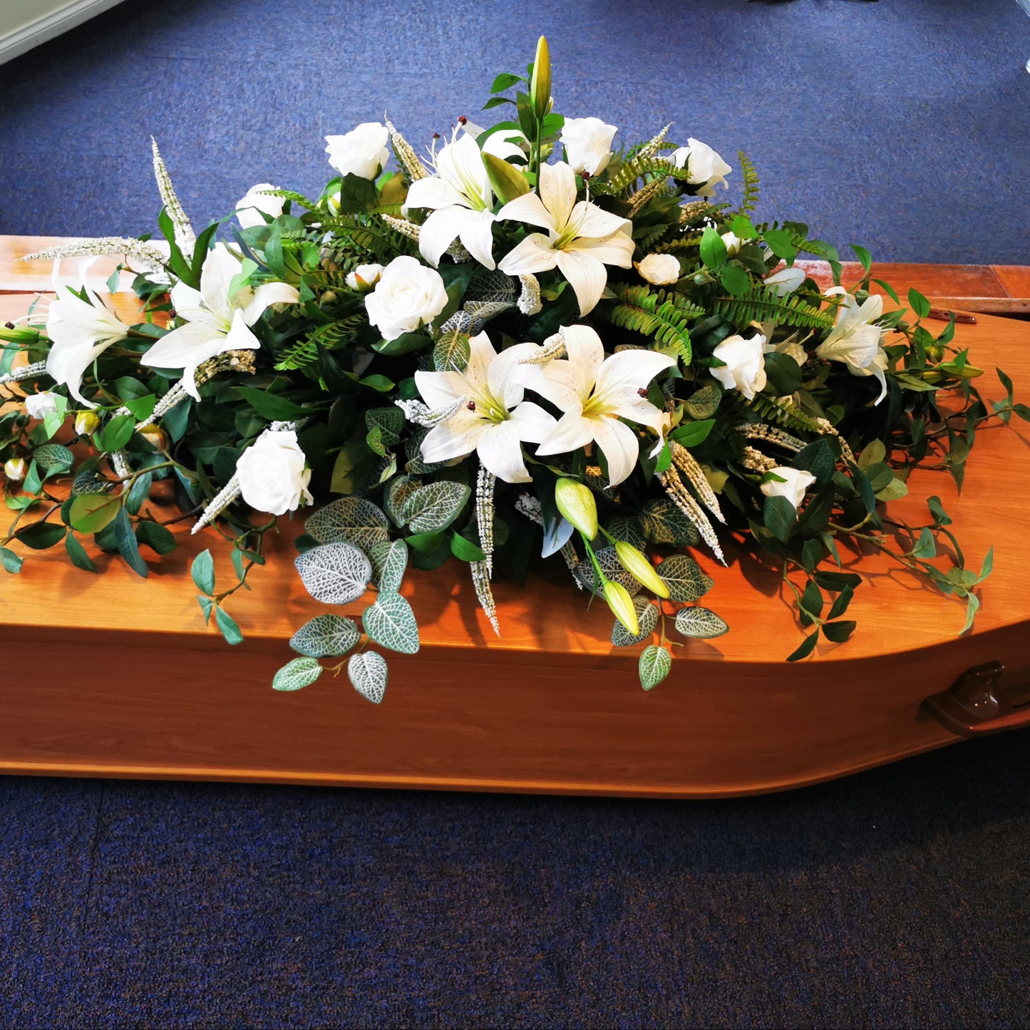 An artificial floral arrangement of white lilies and green foliage displayed on a polished wooden casket, set on a blue carpeted floor.