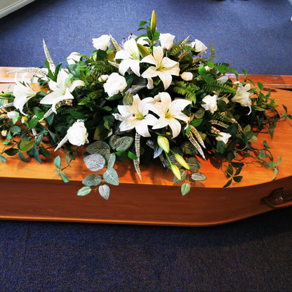 An artificial floral arrangement of white lilies and green foliage displayed on a polished wooden casket, set on a blue carpeted floor.