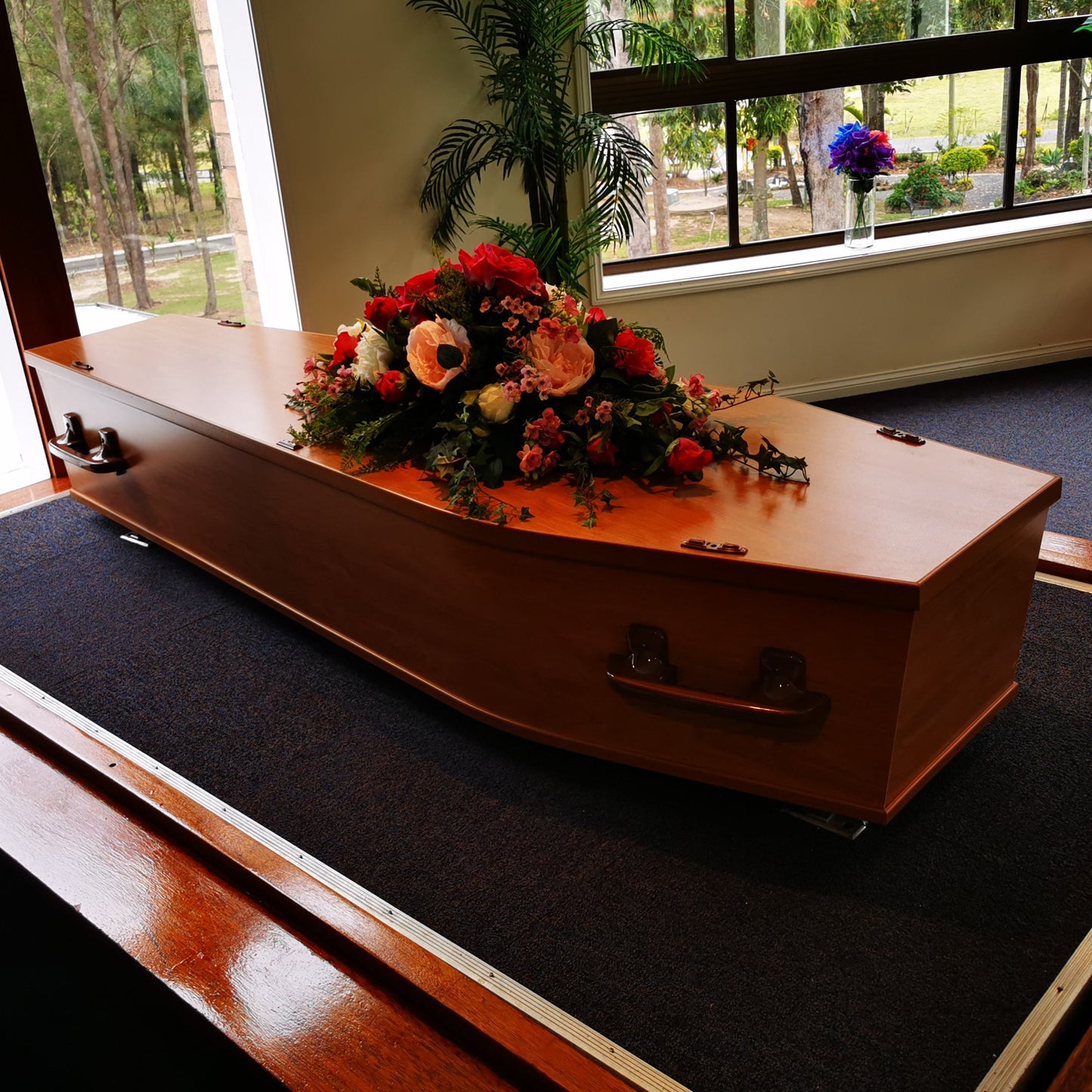 A polished wooden casket with a floral arrangement on top, displayed indoors on a dark carpeted platform near large windows.